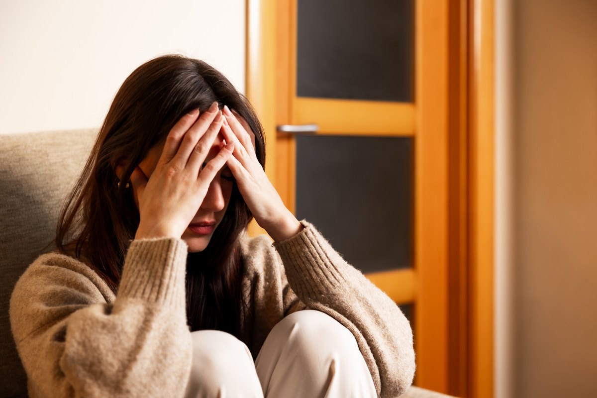 Donna con lunghi capelli castani e maglione beige seduta su un divano, mani sulla fronte e occhi chiusi, porta in legno dietro